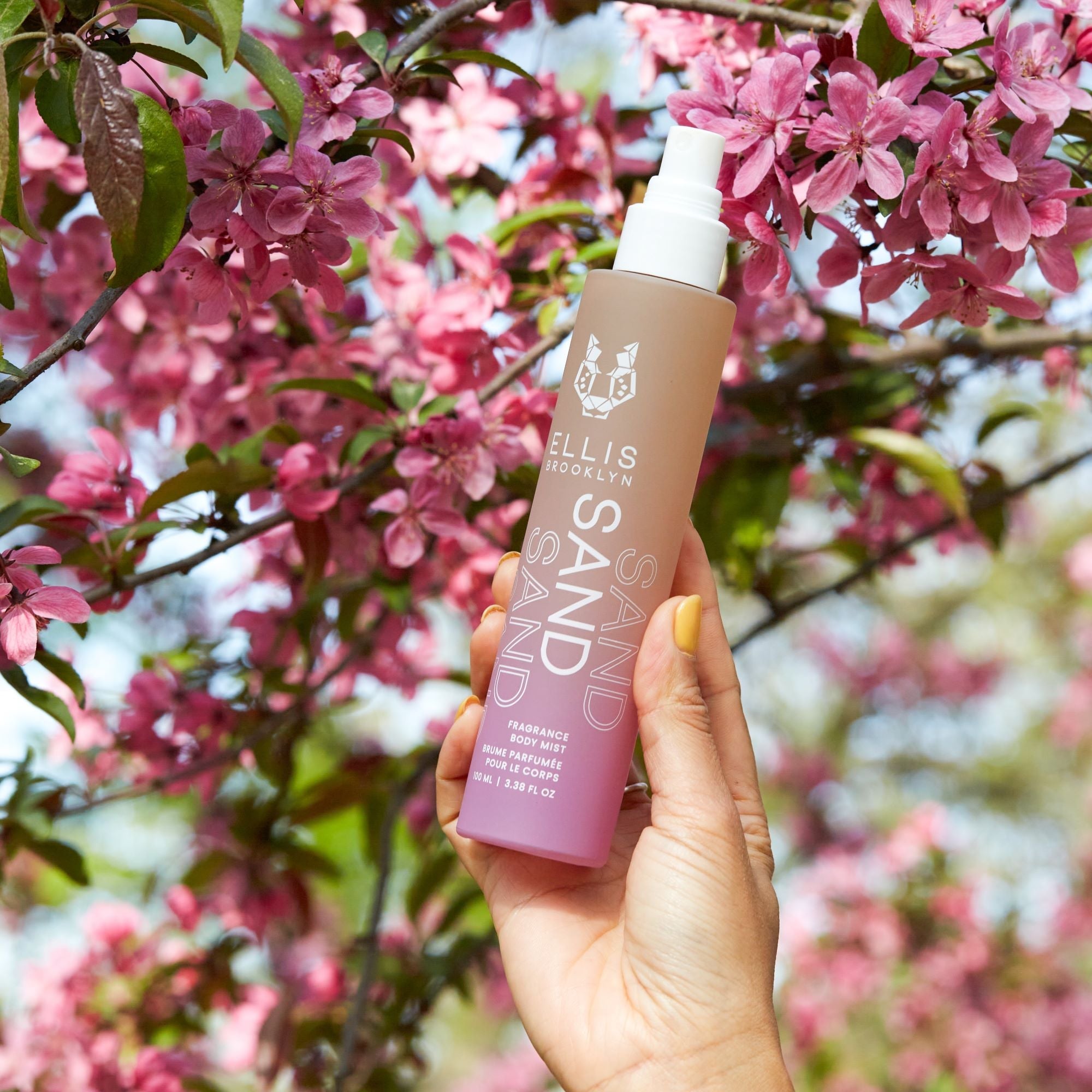 IMAGE 5: MODEL HOLDING SAND FRAGRANCE AND BODY MIST 100ML NEAR FLOWERS