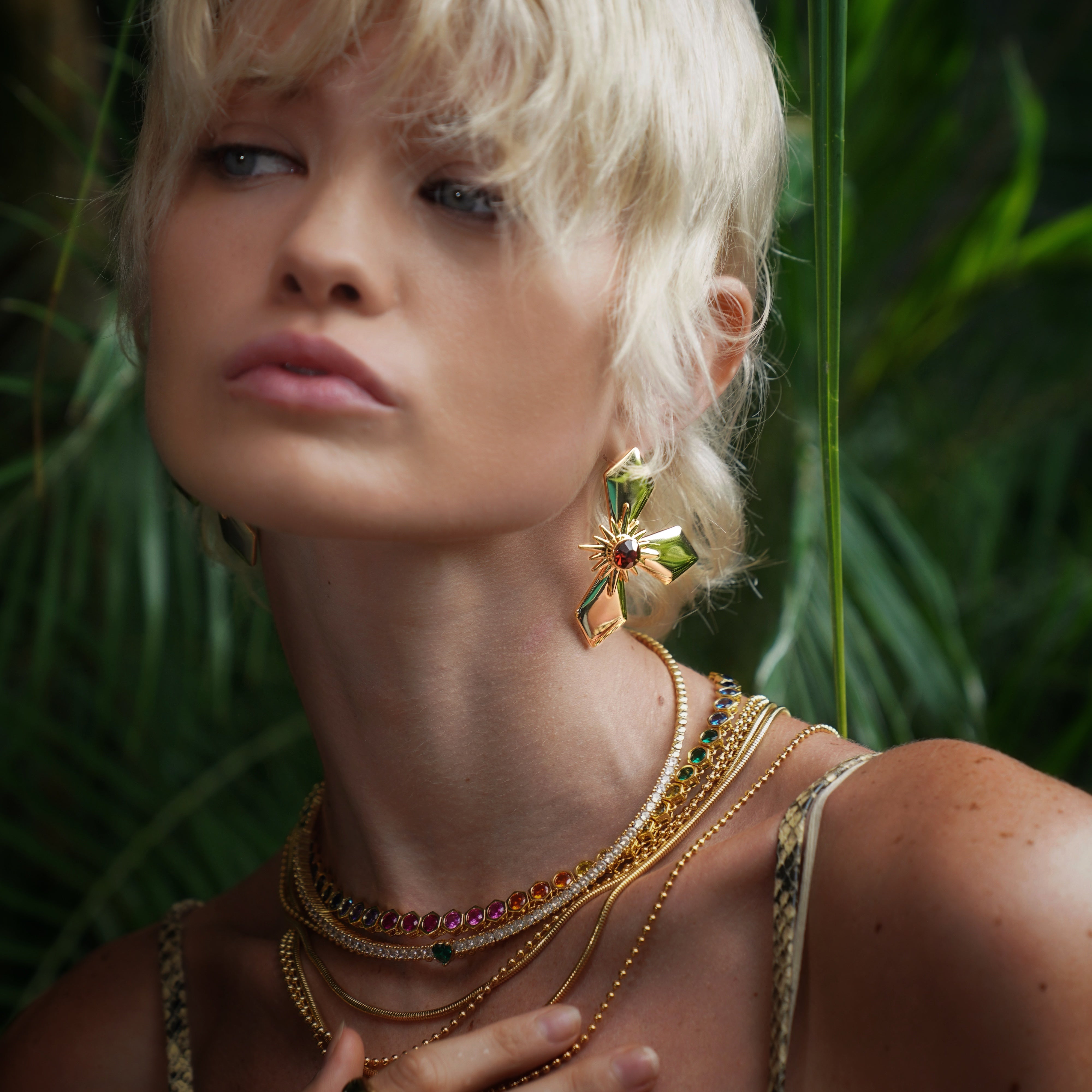 Woman with blonde hair wearing gold jewelry and large earrings in a forest setting.