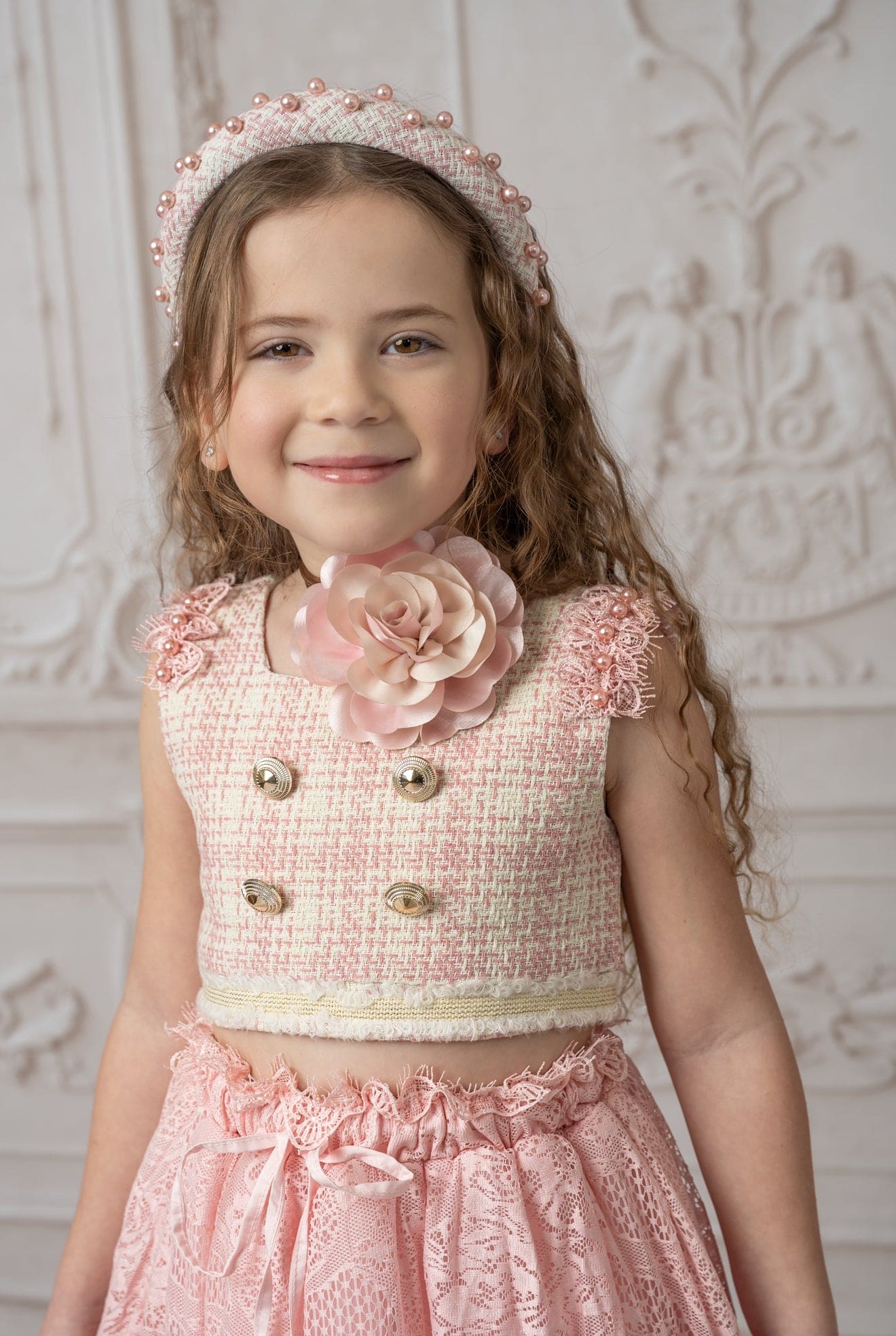 Close up image of girl wearing beige and pink tweed top with gold buttons and a floral necklace paired with a matching tweed headband