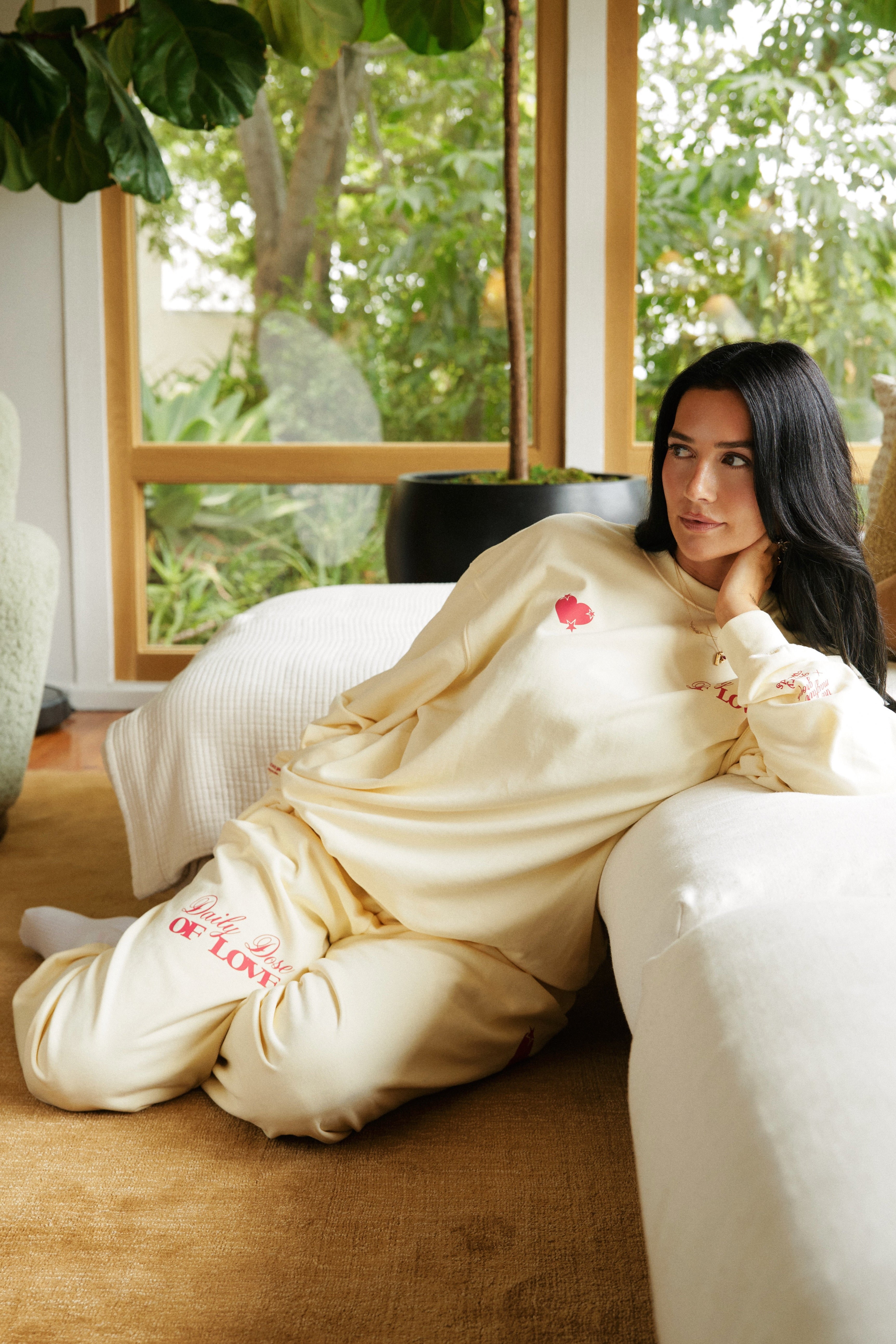Woman in a buttercream-colored outfit with text sitting on a couch in a room with large windows and greenery.