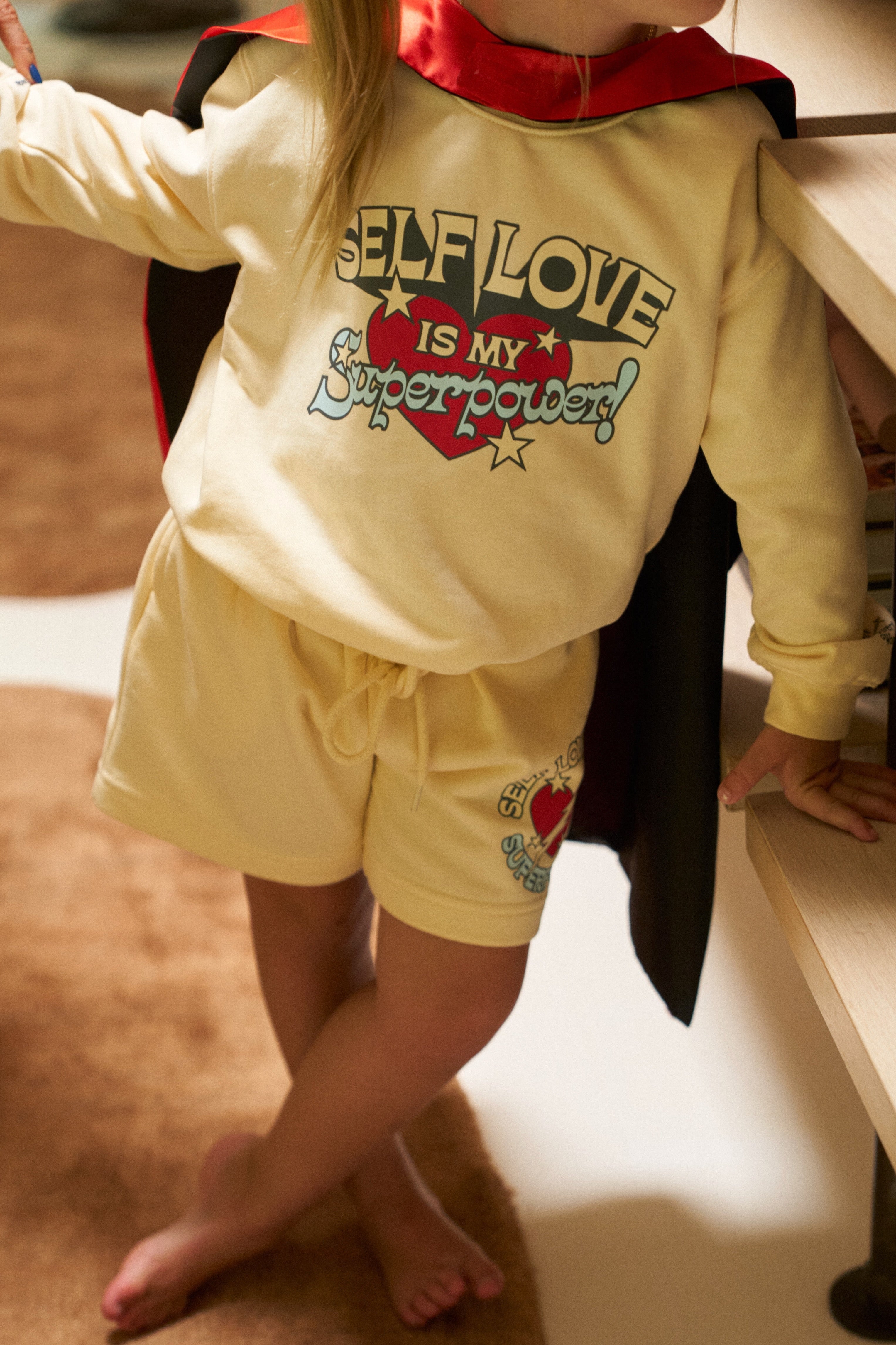 Child wearing a yellow outfit with 'Self love is my superpower' text and a red cape.