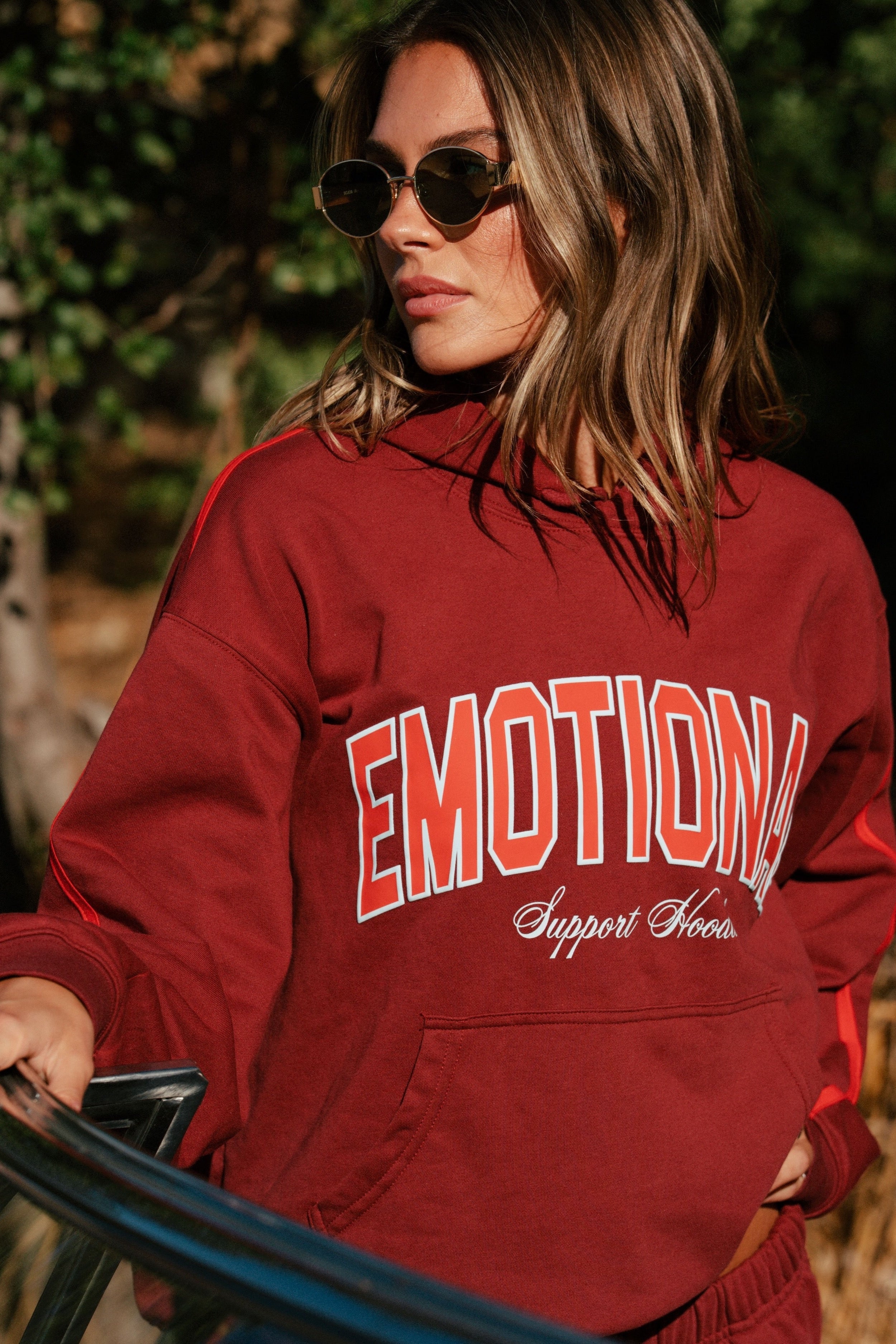 Person wearing a red hoodie with 'EMOTIONAL Support Hoodie' text in a forest setting