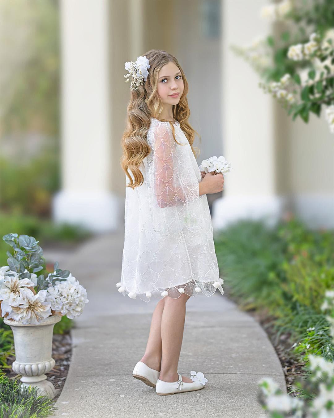 Young girl in a white dress standing outdoors with floral decorations.